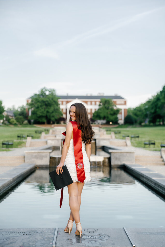 University of Maryland College Park graduate standing on the fountain ledge on McKeldin mall during a sunrise photoshoot. Graduate is facing McKeldin Library, but looking over her left shoulder at the camera while she holds her cap down to her side in her left hand. Her stole is draped over her left shoulder with the UMD logo facing the camera.