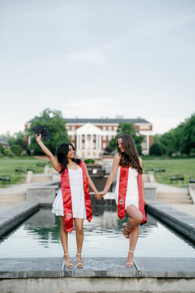 2 graduates striking a cute pose at McKeldin Mall throwing their hands in the air with their graduation cap and popping a leg up a few inches off of the ground captured by UMD Graduation photographer