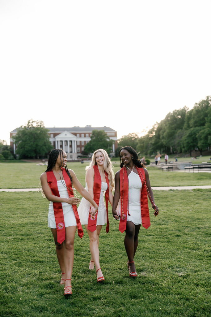 3 best friends smiling as they hold hands walking towards the camera on McKeldin mall at the University of Maryland captured by Brenna Marie Photography