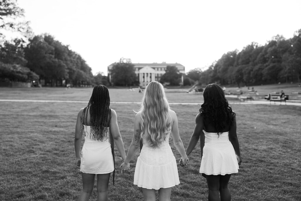 Black and white image of 3 best friends holding hands with their backs to the camera as they walk towards McKeldin Library on McKeldin mall at the University of Maryland captured by Brenna Marie Photography