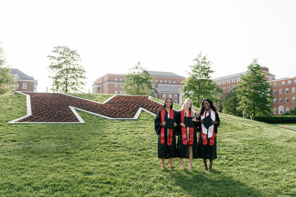 # girls standing in front of "The M," the red flowerbed at the University of Maryland, wearing their graduation gowns and stoles, and holding their caps smiling at the camera captured by UMD Graduation Photographer Brenna Marie Photography
