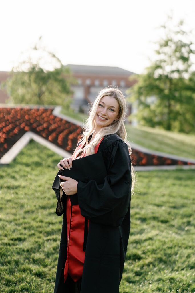 UMD Graduation Photography captured by Brenna Marie Photo. Student posing in front of the flowerbed M wearing her gown and stole while holding her graduation cap in her arms