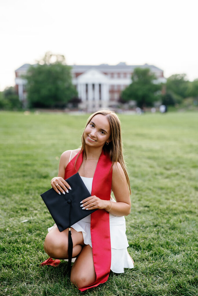 UMD Graduation Photo girl kneeling on grass at McKeldin Mall holding her graduation cap smiling at the camera