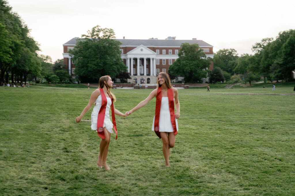 Motion blur slow shutter speed image of 2 best friends running through a field wearing white dresses and their graduation stoles at The University of Maryland McKeldin Mall