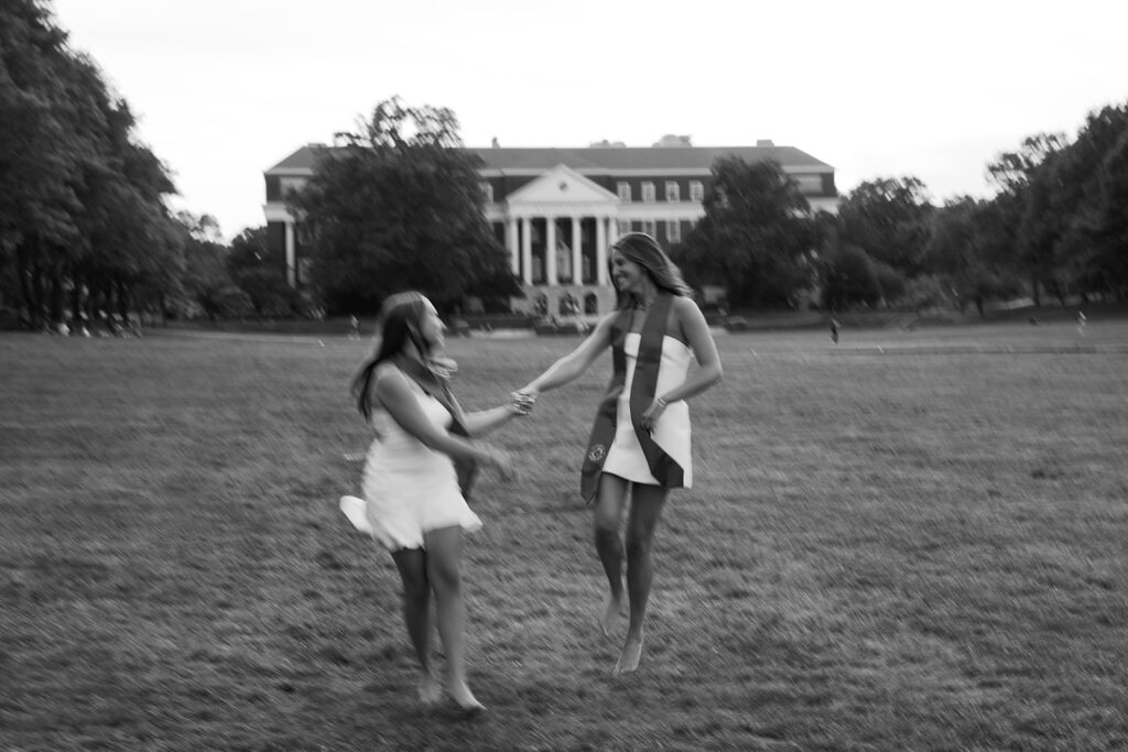 Motion blur slow shutter speed image of 2 best friends running through a field wearing white dresses and their graduation stoles at The University of Maryland McKeldin Mall