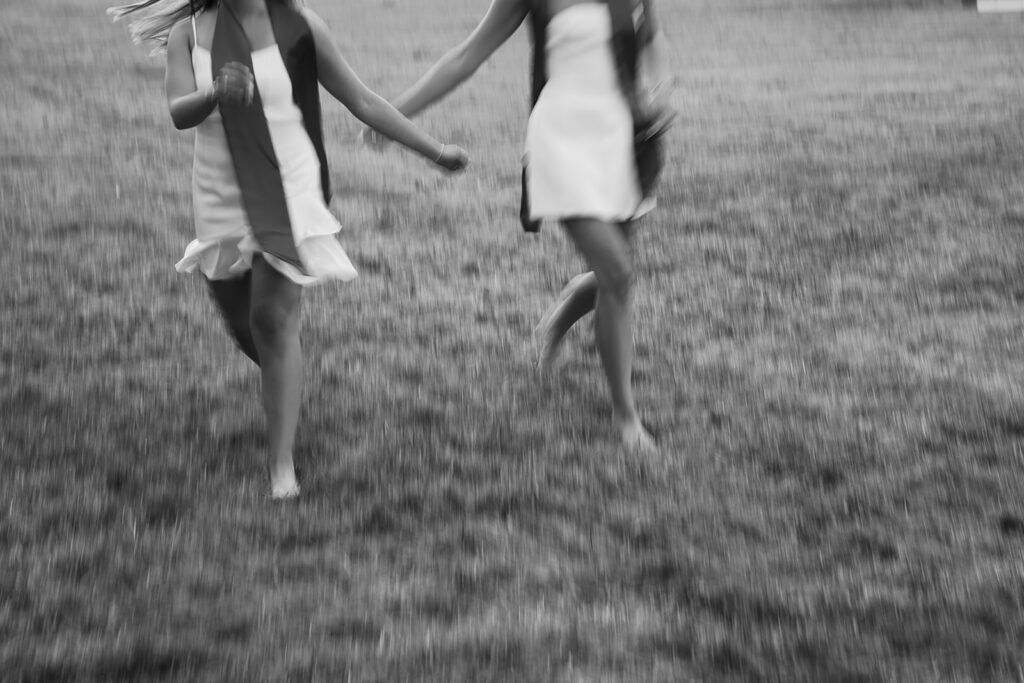 Motion blur slow shutter speed image of 2 best friends running through a field wearing white dresses and their graduation stoles