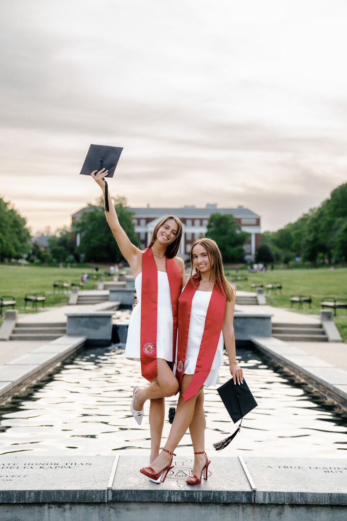 University of Maryland graduates standing on the fountain at McKeldin mall holding their graduation caps and smiling at the camera