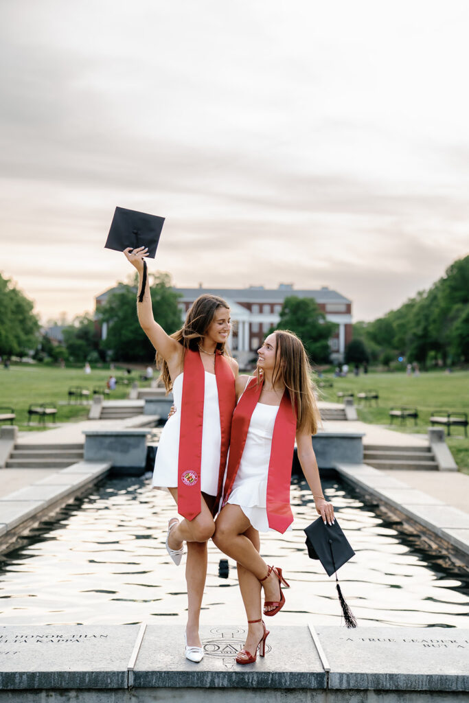 University of Maryland graduates standing on the fountain at McKeldin mall holding their graduation caps and laughing with each other