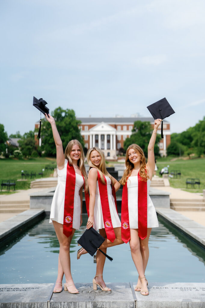 University of Maryland graduates standing on the fountain at McKeldin mall holding their graduation caps and smiling at the camera